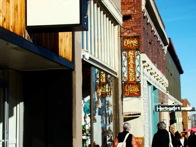 Street view showing storefronts on a sunny day, including a sign for Olympic Standing Glass and Harbor Art, with pedestrians walking.