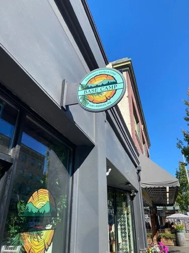 Outdoor sign for Base Camp restaurant with mountain and sunset logo, storefront with windows and awning, clear blue sky.