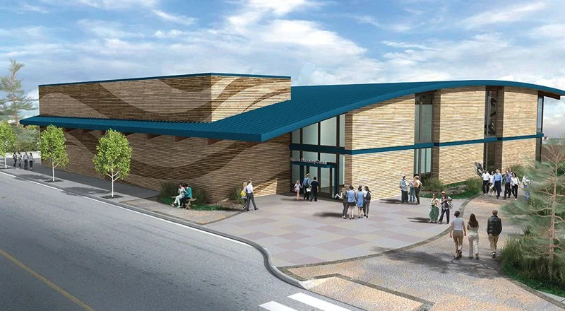 A modern building with a curved blue roof, brick walls, large glass windows, and a glass entrance labeled 'Discovery School'. People are walking around the building on a sidewalk and parking lot area, with a tree-lined street in the foreground.