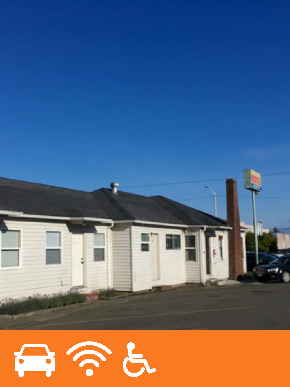 A small white commercial building or restaurant with a black roof, located on a parking lot under a blue sky. There is a tall brick chimney and a sign in the background.