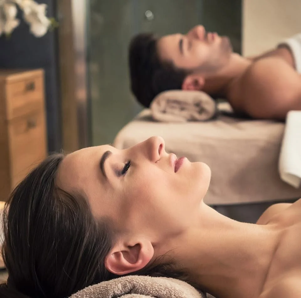 Two people resting on massage tables with beige towels inside a spa room.