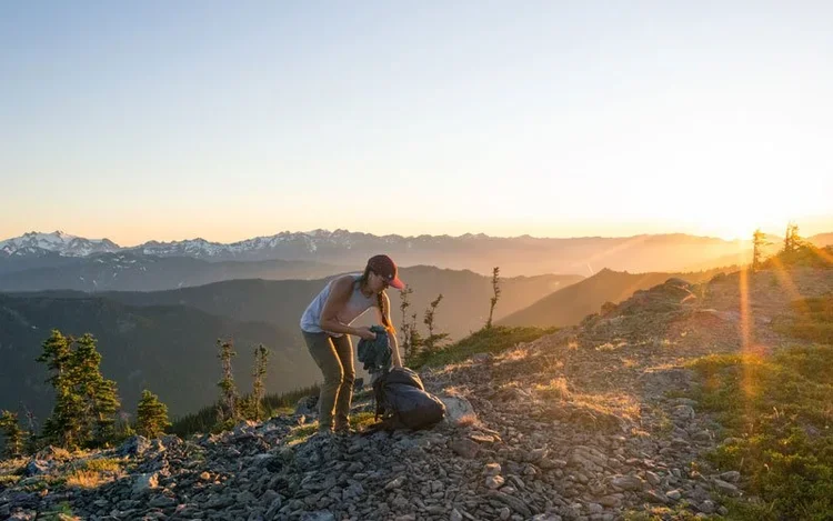 A person gathering belongings on a mountain trail at sunset, with mountain ranges in the background and a backpack nearby.