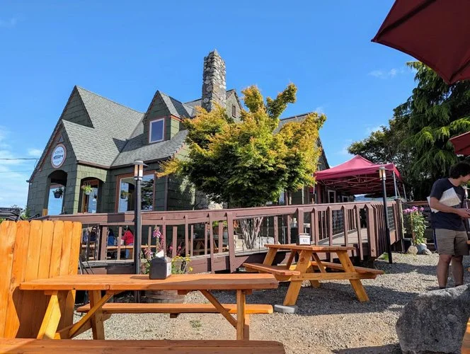 A rustic, dark green house with steep rooflines and a stone chimney, surrounded by outdoor seating with picnic tables, umbrellas, and people, under a clear blue sky.