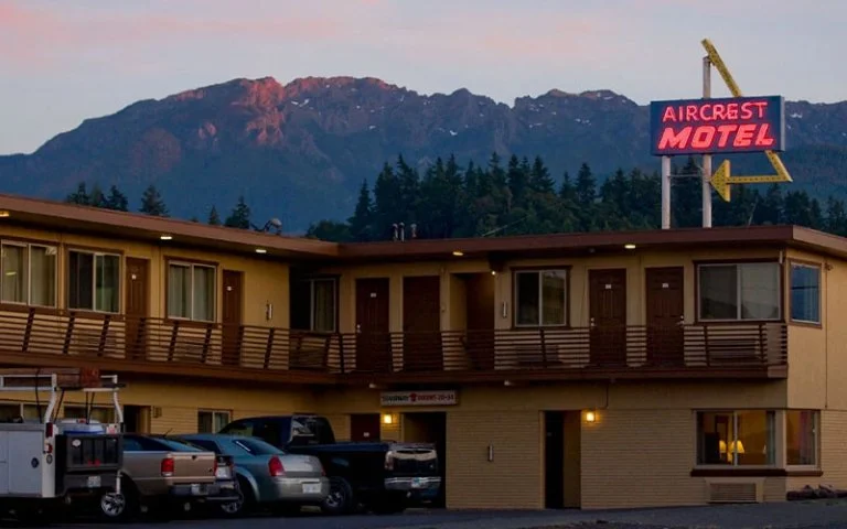 Two-story motel called Aircrest Motel with a parking lot in front, mountain range in the background, and a neon sign with an arrow pointing left.