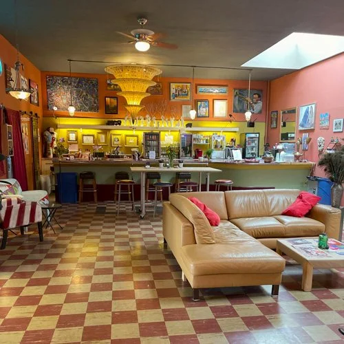 Interior of a cozy cafe with a bar counter, bar stools, a beige sectional sofa with red pillows, a small coffee table, and a patterned floor. Decor includes colorful framed pictures and a large chandelier.