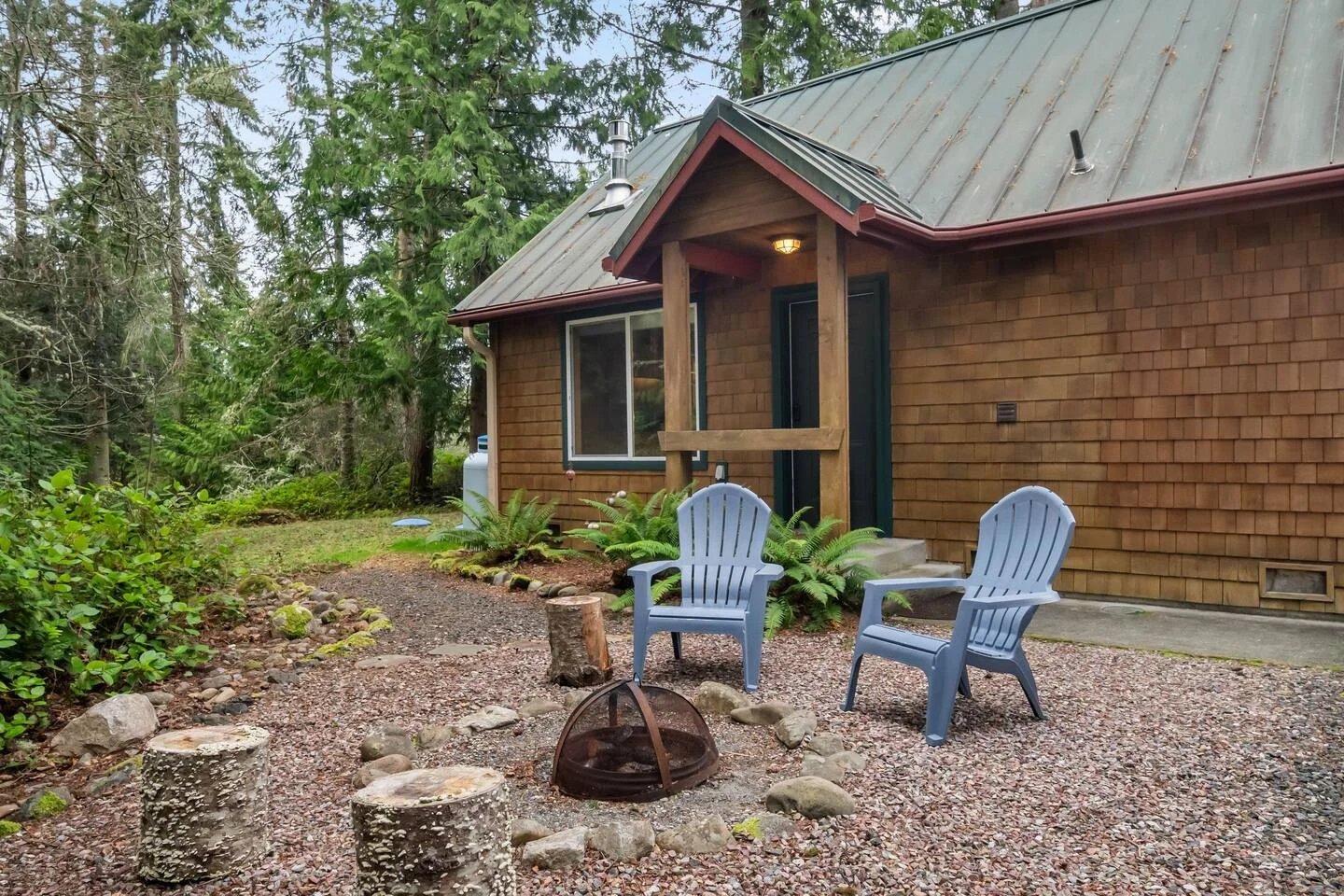 Backyard with two blue plastic chairs around a fire pit, gravel ground, wooden Chinese-style stumps, greenery, and a brown wooden house with a metal roof and a porch.