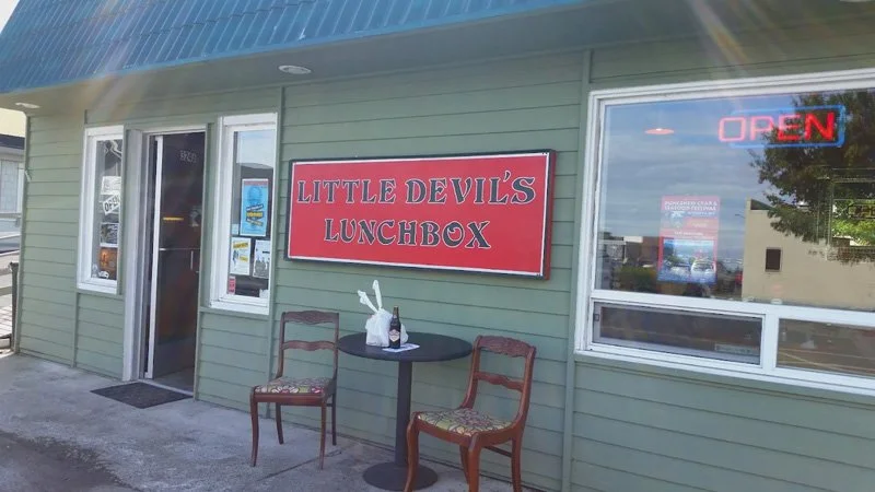 Exterior of Little Devil's Lunchbox restaurant with a red sign, outdoor table with two chairs, and a window with an illuminated 'OPEN' sign.