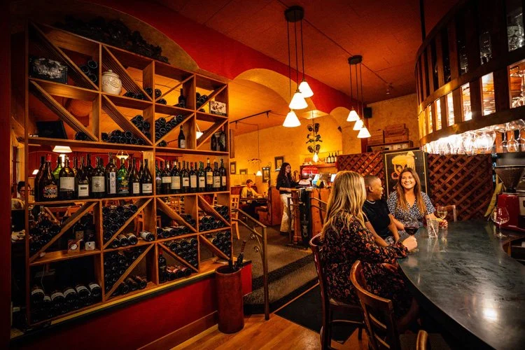 A wine bar with a large wine rack filled with bottles, a bar counter where three women are seated chatting, warm lighting, and a bartender in the background.