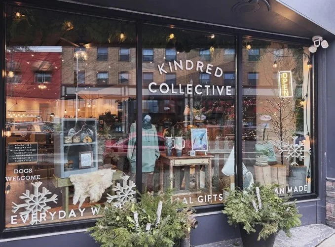 Storefront window of Kindred Collective with holiday decorations, snowflake decals, and potted greenery outside.