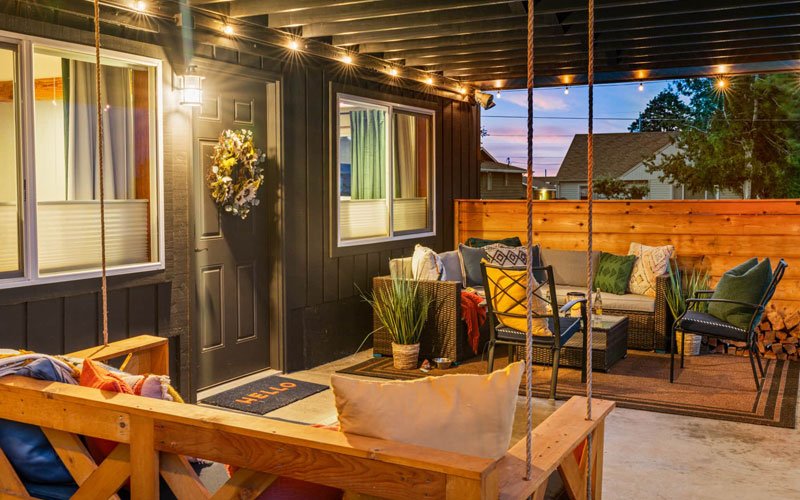 A cozy outdoor patio with wicker and fabric chairs, plants, a rug, string lights, a wooden privacy fence, and a door with a wreath, set against a twilight sky.