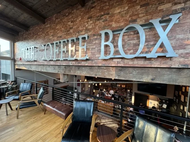 Interior of a coffee shop named 'The Coffee Box,' with a brick wall, large blue letters spelling out the name, and various chairs and tables for seating.