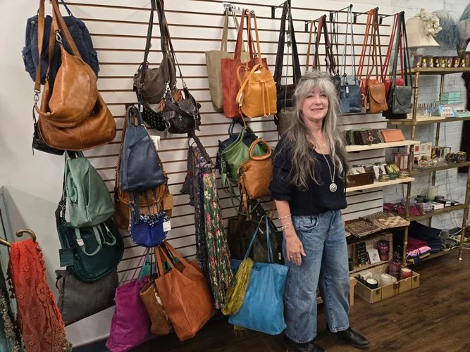 A woman with long gray hair, wearing a black top, jeans, and black shoes, standing in front of a display of handbags on a slat wall in a store. The handbags are in various colors and styles, hanging from hooks. To the right, shelves hold miscellaneous items, including jewelry and decorative boxes.