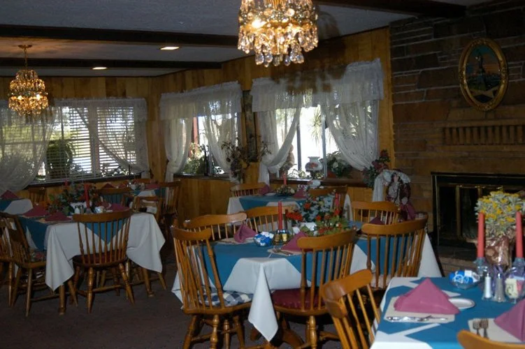 A decorated dining room with tables covered in white and blue tablecloths, set with pink napkins and floral centerpieces, surrounded by wooden chairs, with large windows, lace curtains, chandeliers, and a brick fireplace.