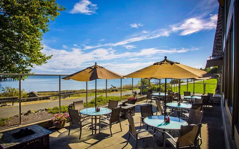 Outdoor patio with round tables, chairs, and large umbrellas overlooking a lake under a partly cloudy sky.