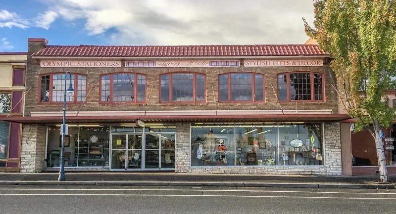 Two-story brick building with large front windows on the ground floor and smaller arched windows on the second floor, with signs for Olympic Stationers and Stylish Gents & Decor, on a street with a lamppost and trees.