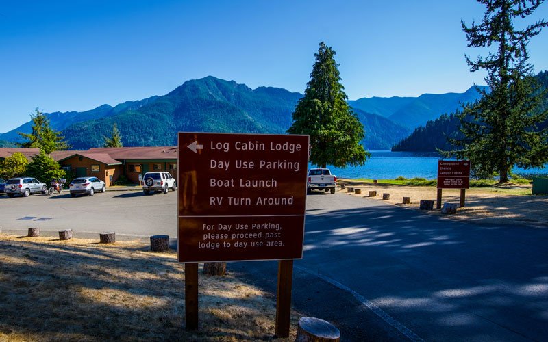 Sign at a lakeside parking lot pointing to log cabin lodge, day use parking, boat launch, and RV turn around with mountains and trees in the background.