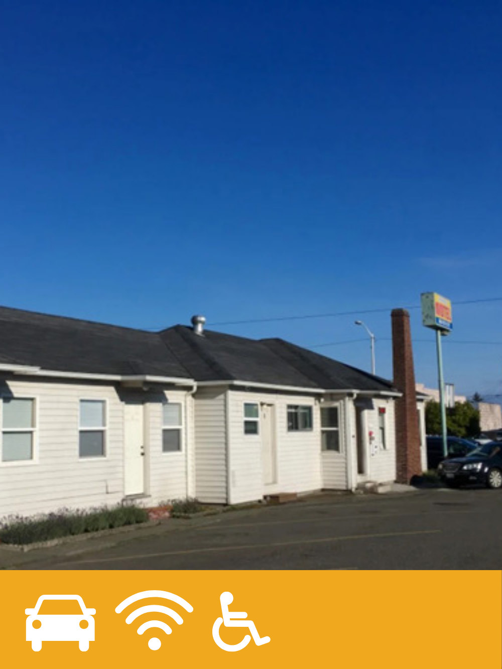 A small white commercial building or restaurant with a black roof, located on a parking lot under a blue sky. There is a tall brick chimney and a sign in the background.