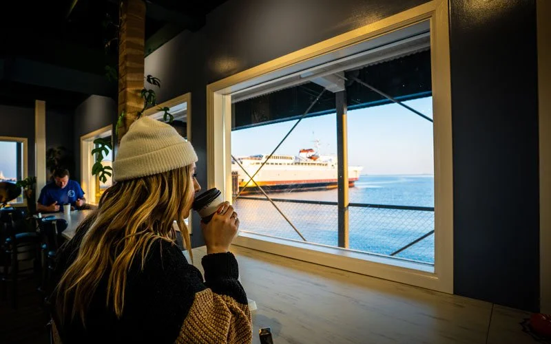 A woman with long blonde hair wearing a white knit hat and black sweater, drinking coffee while sitting at a seaside café, looking out a window at a ferry on the water.
