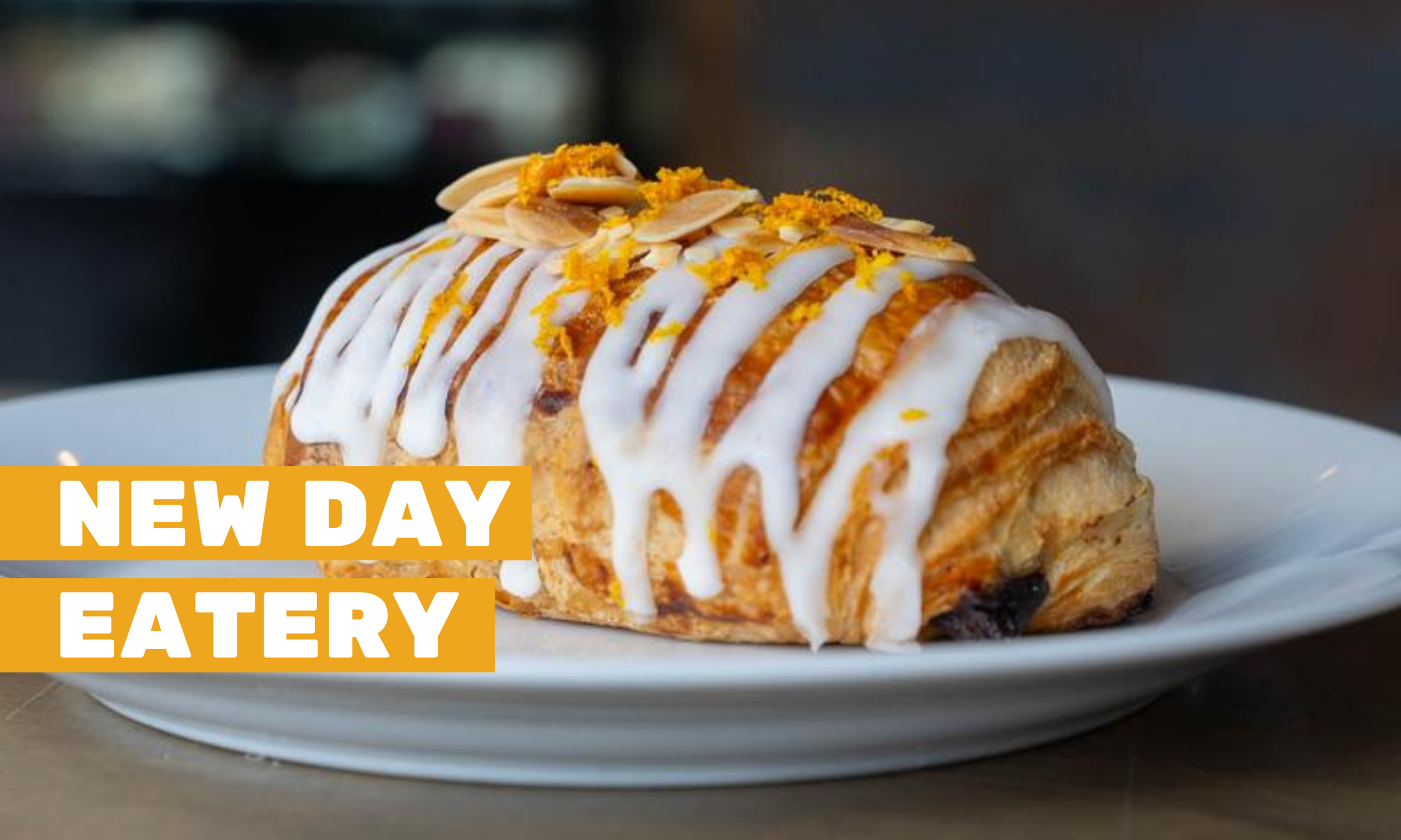 A citrus, almond, frosted scone on a white plate with text on the photo that reads "New Day Eatery" on it.