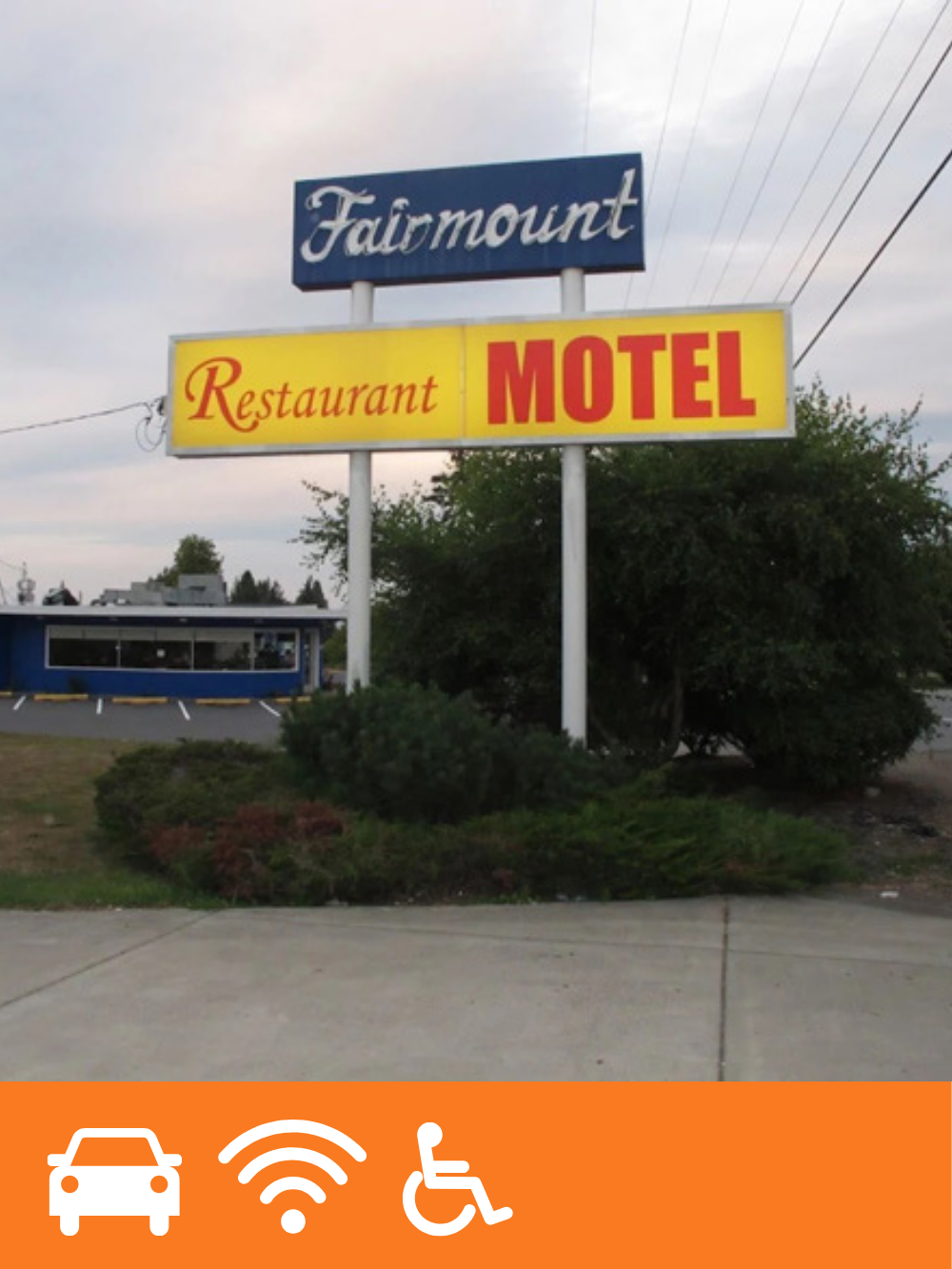 Signboard for a motel with a restaurant, located in a parking lot with bushes in front and a building in the background.