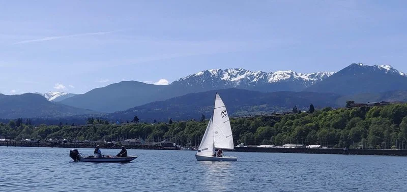 A fishing boat and a sailboat on a lake, with green hills and snow-capped mountains in the background under a clear sky.