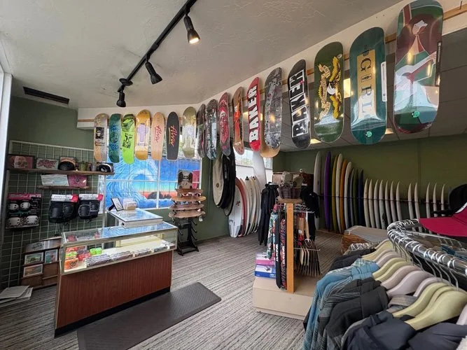 Skateboards hanging on the wall in a skate shop with surfboards, apparel, and accessories displayed inside.