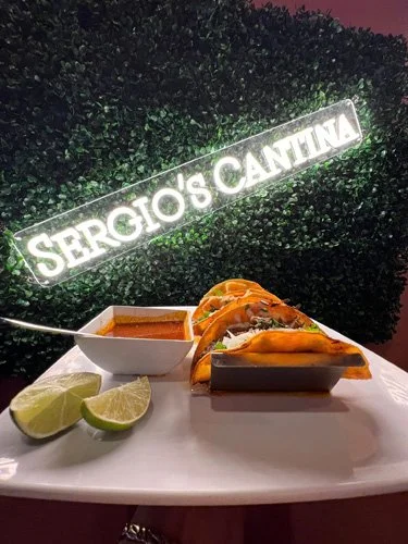 A table with two tacos, lime wedges, and a small bowl of sauce in front of a hedge with a sign that reads 'Sergio's Cantina'.