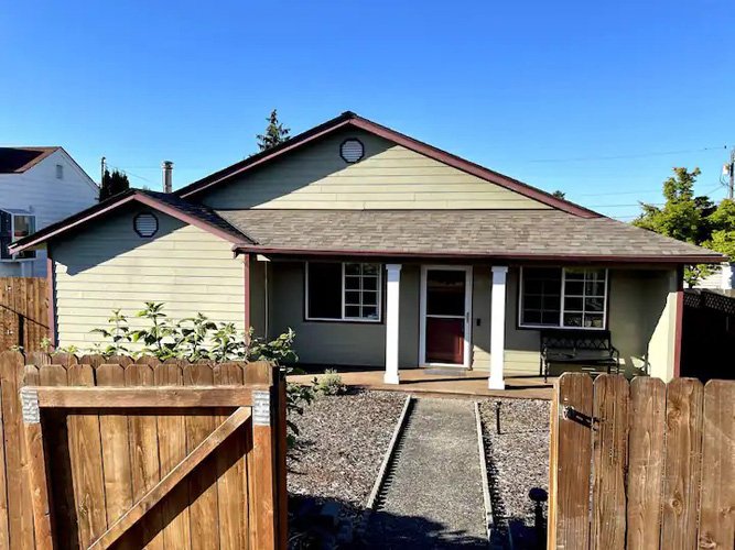 A small house with a green exterior, a gabled roof, and a covered porch with white columns. Railroad tracks run through the front yard, leading to the porch. A wooden fence encloses the yard.