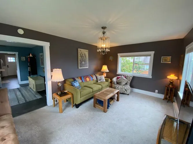 Living room with dark gray walls, a green sofa, a patterned armchair, two lamps, a chandelier, a coffee table, and a window with a view of trees.