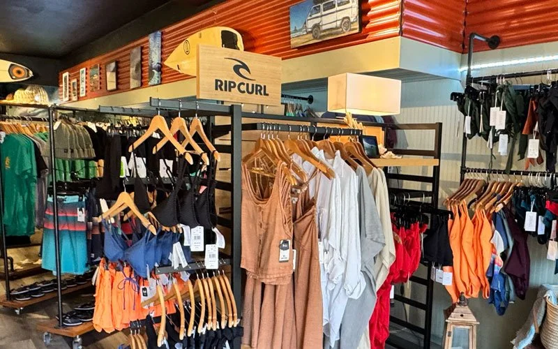 Clothing display inside a retail store, featuring swimsuits, tank tops, and beach accessories, with a Rip Curl surfboard and a small photo of a vehicle on the wall.