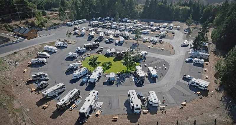 A large parking lot filled with numerous RVs and camper trailers, arranged in a circular pattern around a small green area with trees, surrounded by a wooded area.