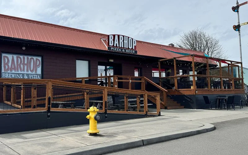 Exterior of a restaurant named Barhop offering pizza and beer, with a large outdoor deck area with seating, a red metal roof, and a yellow fire hydrant in front.