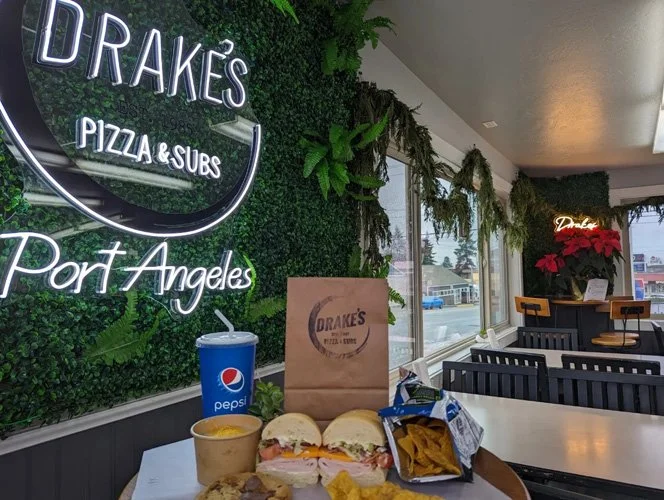 Inside of Drake's Pizza & Subs restaurant in Port Angeles with a neon sign, green wall decor, and a fast food meal in the foreground that includes a soda, sandwich, chips, and dip.