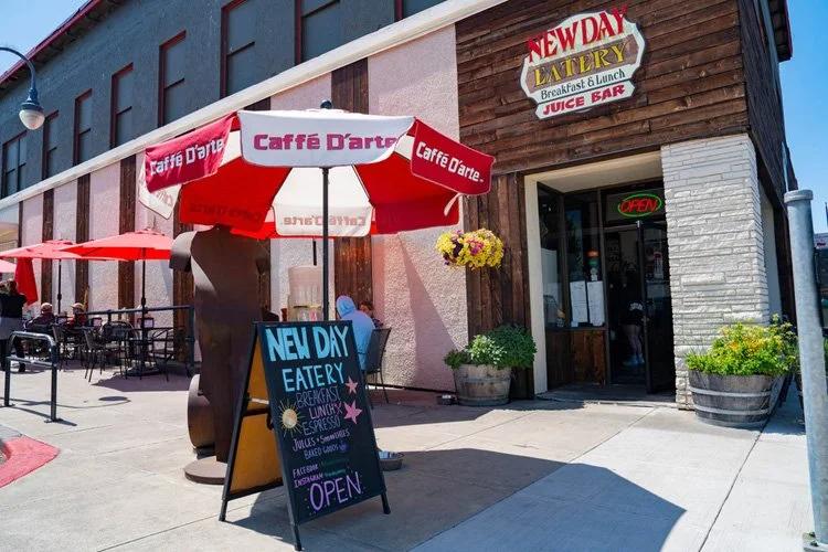 The exterior of a restaurant called New Day Eatery with outdoor seating, a red and white umbrella, a chalkboard sign, and a flower pot outside the entrance.