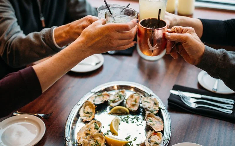 People clinking glasses over a table with a platter of oysters and lemon wedges.