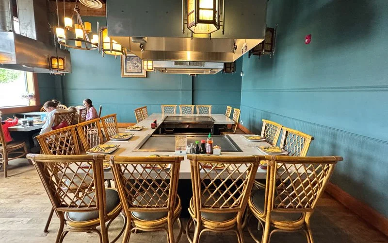 Empty round dining table with a built-in teppanyaki grill in a restaurant, surrounded by wooden lattice-backed chairs, with condiment bottles and set plates, in a room with teal walls, large windows, and decorative hanging lights.