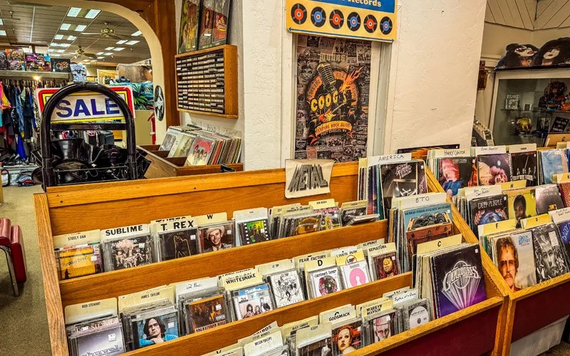 View of a record store display with vinyl albums and CDs, including a sale sign in the background, interior of the store with racks and shelves of music, and colorful posters on the walls.