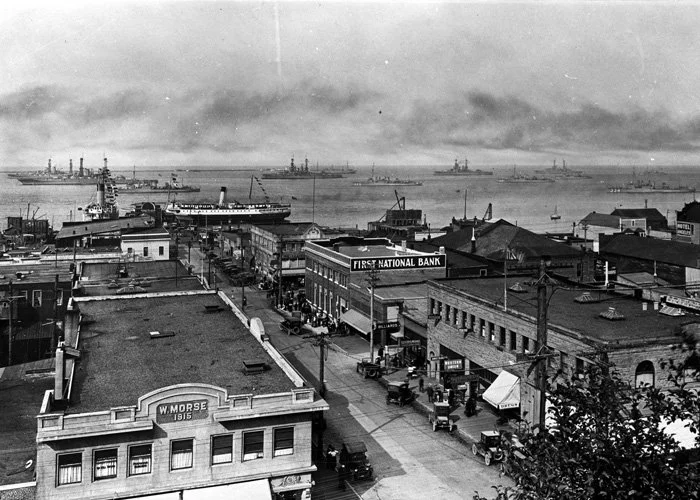 Historical black-and-white photograph of a city waterfront with ships in the harbor, a busy street with vintage automobiles, and buildings including a bank and a sign reading 'W. Morse 1915'.