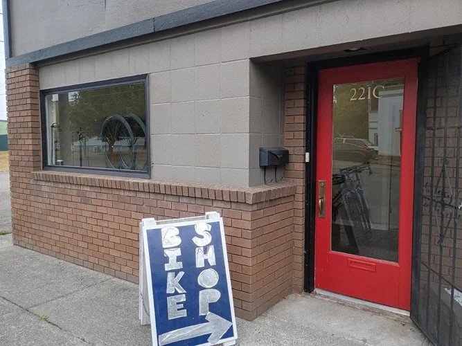 A sidewalk scene showing a red door at a brick and beige building. A sign with a blue background and white paint offers bike rentals, with an arrow pointing to the right.