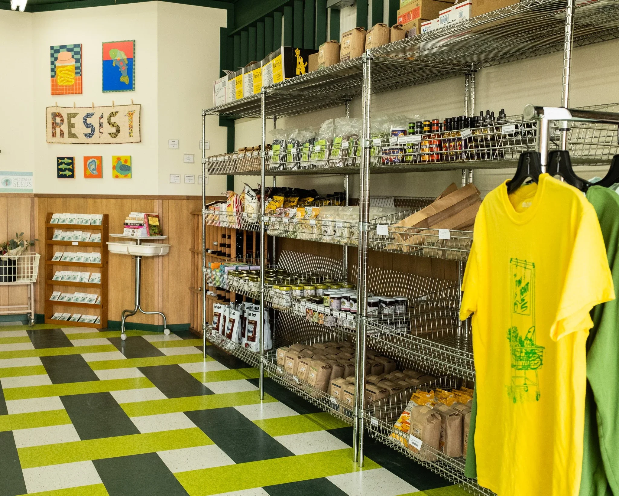 Interior of a small store or café with metal shelves stocked with packaged food and snacks, and a clothing rack with yellow and green t-shirts, alongside a patterned green, black, and white floor.