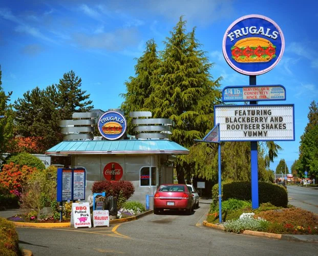 Frugal's fast food restaurant with a large circular sign and a marquee advertising blackberry and root beer shakes. A red car is parked in front of the building, surrounded by trees and bushes.