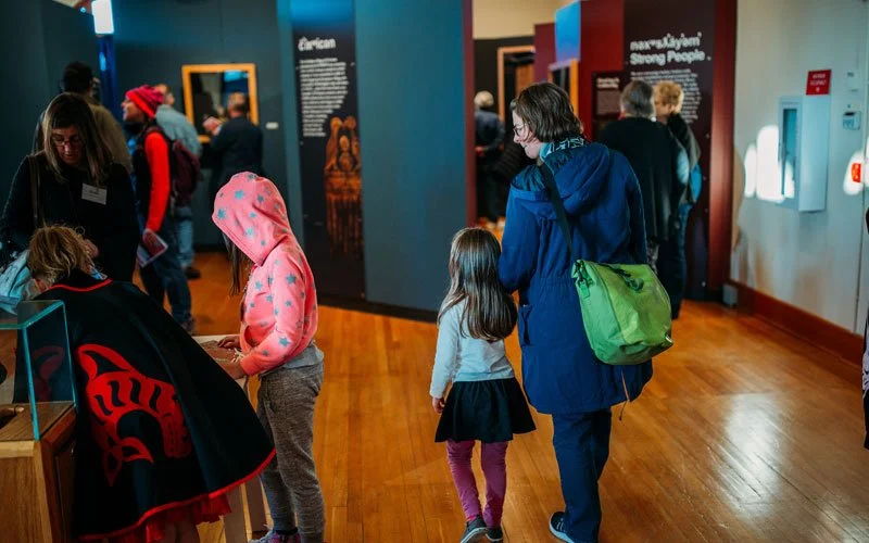 Visitors, including children and adults, explore exhibits at a museum with wooden floors and informational displays on the walls.