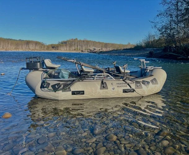 Inflatable boat on a rocky riverbank with oars, fishing gear, and seating, with a river and trees in the background
