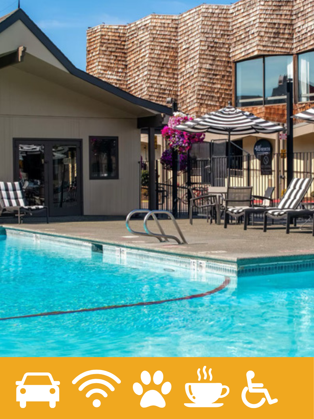 A swimming pool area next to a building with a sloped roof and wooden shingles. There are striped lounge chairs, a table with chairs, and umbrellas, with pink flowers hanging from a pole. The sky is blue with some clouds.