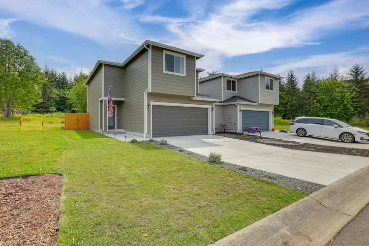 A modern, two-story gray house with attached garages, surrounded by a lawn and trees, with a driveway and a parked car.