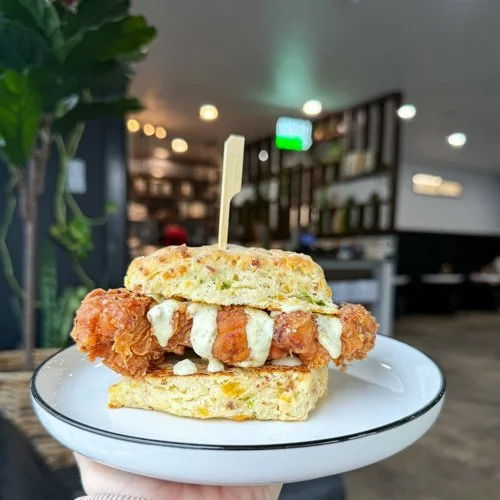 Fried chicken sandwich with creamy sauce on a bun, served on a white plate with a fork, inside a modern restaurant