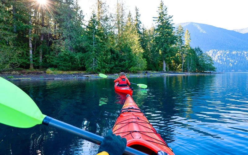 Person kayaking on a calm, clear lake surrounded by tall trees and mountains in the distance, with sunlight peeking through the forest.