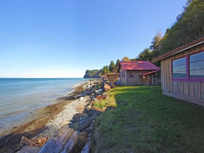 Beachside houses with red roofs and large windows, green grass, rocky shoreline, clear blue sky, and trees in the background.