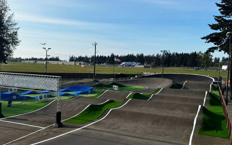 An outdoor skatepark with various ramps and jumps, surrounded by grassy areas and trees, with an airplane flying in the sky in the background.