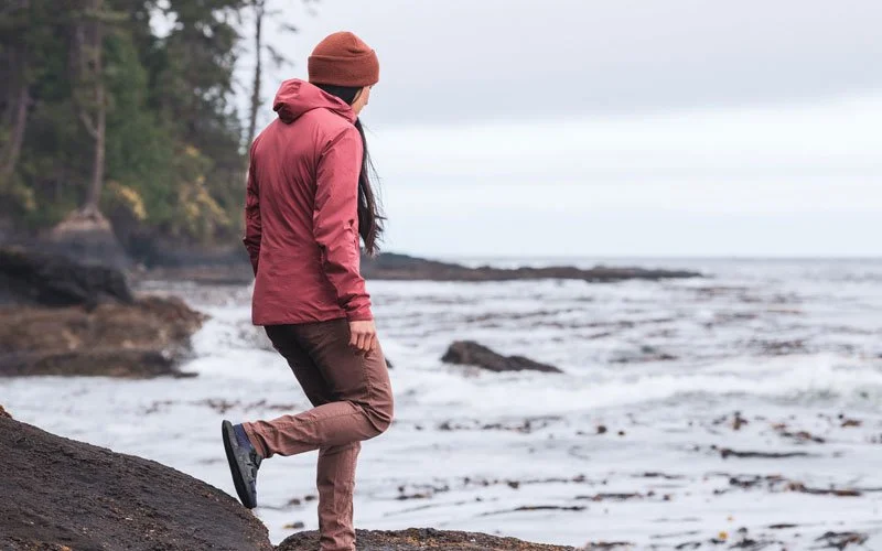 A person stands on a rocky shoreline looking out at the water, wearing a red jacket, brown pants, and a beanie cap.